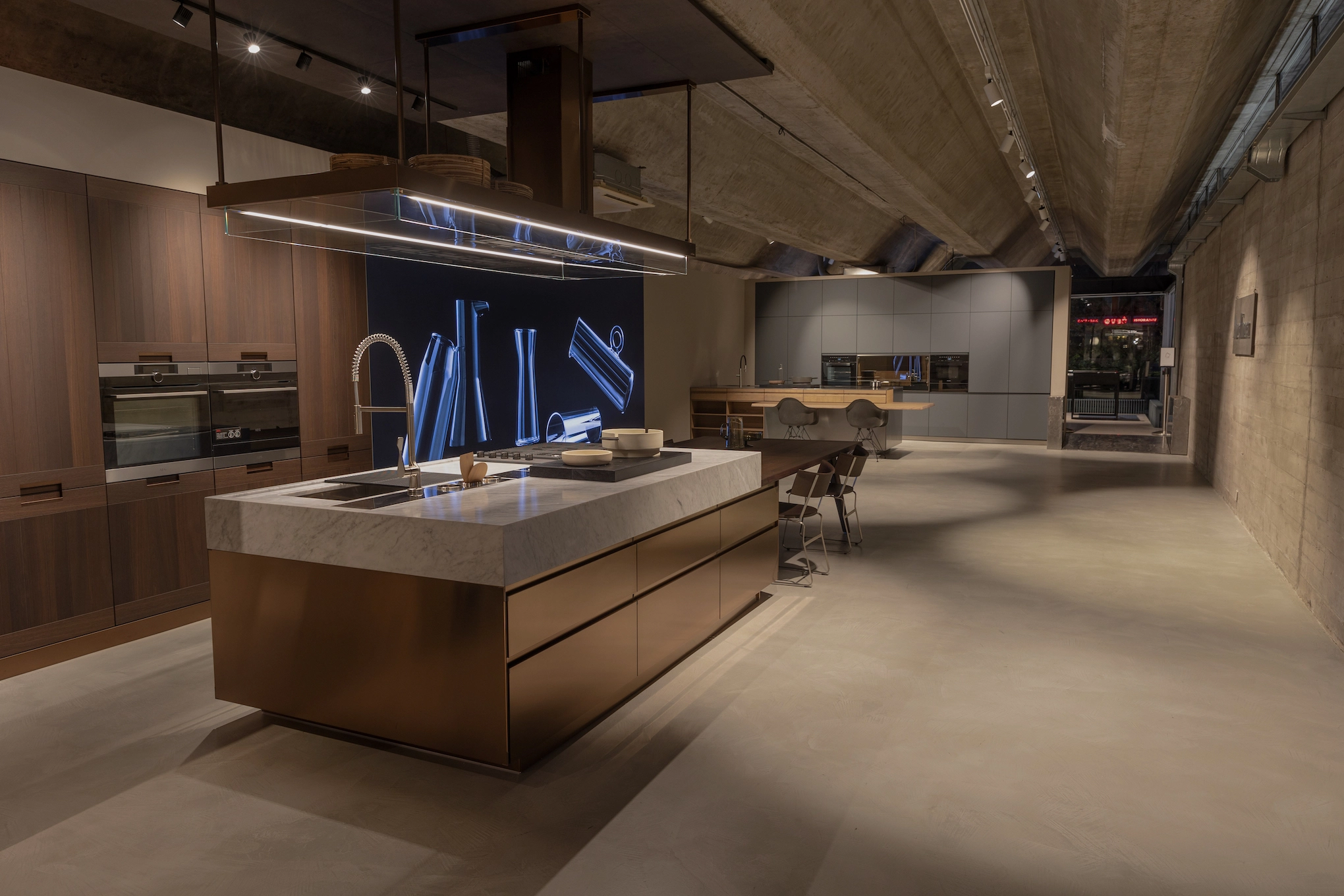 Photograph of the interior of the Arclinea showroom in Fontana Costabissara. Two kitchens are visible, one with a copper-colored steel island and one with light gray columns.